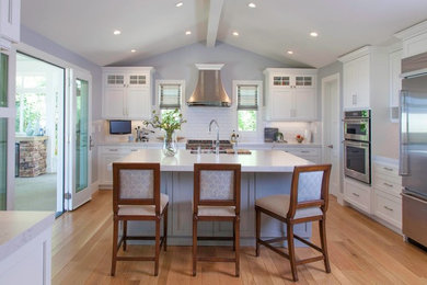 Mid-sized elegant l-shaped medium tone wood floor and beige floor open concept kitchen photo in San Diego with a farmhouse sink, recessed-panel cabinets, white cabinets, quartz countertops, white backsplash, subway tile backsplash, stainless steel appliances, an island and white countertops