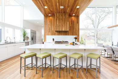 Large contemporary l-shaped eat-in kitchen in Cincinnati with flat-panel cabinets, white cabinets, stainless steel appliances, with island, an undermount sink, solid surface benchtops, white splashback, glass sheet splashback, medium hardwood floors, brown floor and grey benchtop.