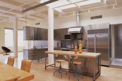Large urban l-shaped light wood floor open concept kitchen photo in New York with flat-panel cabinets, stainless steel cabinets, stainless steel countertops, stainless steel appliances and an island