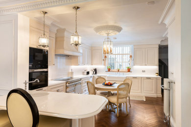 Large victorian l-shaped kitchen/diner in London with a belfast sink, shaker cabinets, beige cabinets, engineered stone countertops, white splashback, engineered quartz splashback, stainless steel appliances, dark hardwood flooring, a breakfast bar, brown floors and white worktops.