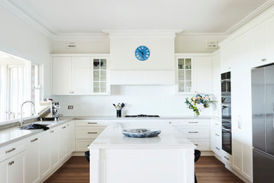 Inspiration for a large traditional u-shaped open plan kitchen in Sydney with a double-bowl sink, recessed-panel cabinets, white cabinets, quartz benchtops, white splashback, cement tile splashback, black appliances, dark hardwood floors, with island, brown floor and white benchtop.