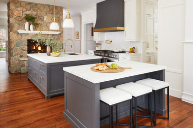 Large traditional u-shaped open plan kitchen in New York with a farmhouse sink, shaker cabinets, white cabinets, marble benchtops, white splashback, marble splashback, stainless steel appliances, medium hardwood floors, multiple islands, brown floor and white benchtop.