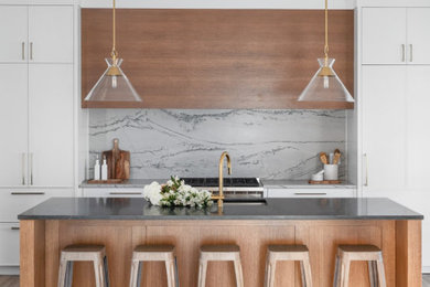 Example of a large trendy medium tone wood floor and tray ceiling kitchen design in Calgary with white cabinets, gray backsplash, stone slab backsplash, stainless steel appliances, an island, black countertops and flat-panel cabinets