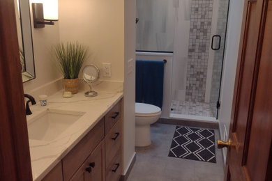 Mid-sized transitional bathroom in Minneapolis with flat-panel cabinets, medium wood cabinets, an open shower, a two-piece toilet, gray tile, ceramic tile, beige walls, vinyl floors, an undermount sink, engineered quartz benchtops, grey floor, a hinged shower door, white benchtops and a built-in vanity.