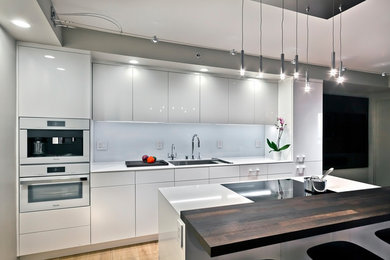 Small single-wall open plan kitchen in Minneapolis with an undermount sink, flat-panel cabinets, white cabinets, quartz benchtops, white splashback, glass sheet splashback, white appliances, medium hardwood floors, with island, brown floor and white benchtop.