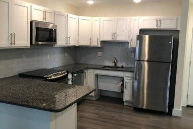 Example of a large minimalist u-shaped dark wood floor and brown floor kitchen pantry design in Orlando with a single-bowl sink, shaker cabinets, white cabinets, gray backsplash, stainless steel appliances, a peninsula and black countertops