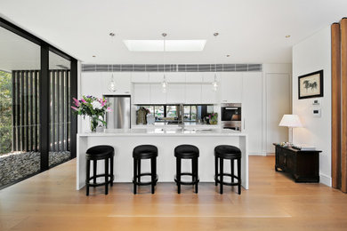 Photo of a large modern single-wall open plan kitchen in Sydney with a double-bowl sink, flat-panel cabinets, white cabinets, marble benchtops, metallic splashback, glass sheet splashback, stainless steel appliances, medium hardwood floors, with island, brown floor and white benchtop.