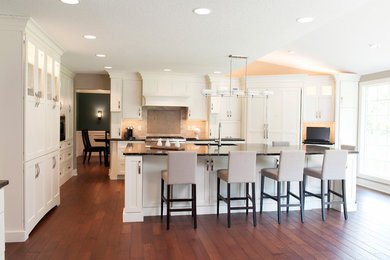 Example of a large transitional l-shaped medium tone wood floor and brown floor eat-in kitchen design in Other with an undermount sink, recessed-panel cabinets, white cabinets, quartz countertops, beige backsplash, limestone backsplash, stainless steel appliances, an island and black countertops