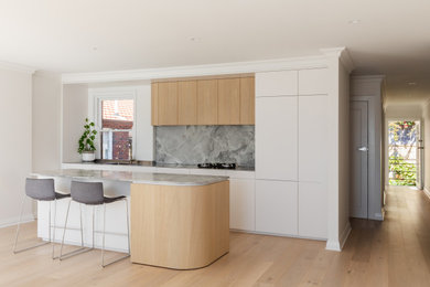Design ideas for a mid-sized contemporary galley open plan kitchen in Sydney with an integrated sink, flat-panel cabinets, light wood cabinets, stainless steel benchtops, grey splashback, stone slab splashback, panelled appliances, light hardwood floors, with island and grey benchtop.