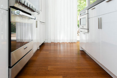 Mid-sized trendy single-wall medium tone wood floor enclosed kitchen photo in Denver with flat-panel cabinets, white cabinets, stainless steel countertops, stainless steel appliances and an island