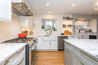 This is an example of a large transitional l-shaped open plan kitchen in Ottawa with a farmhouse sink, shaker cabinets, white cabinets, quartz benchtops, white splashback, ceramic splashback, stainless steel appliances, medium hardwood floors, with island, brown floor and white benchtop.