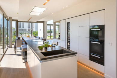 Zweizeilige, Große Moderne Wohnküche mit integriertem Waschbecken, flächenbündigen Schrankfronten, weißen Schränken, Betonarbeitsplatte, schwarzen Elektrogeräten, braunem Holzboden, zwei Kücheninseln und braunem Boden in Köln
