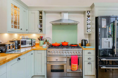 Photo of a medium sized classic cream and black l-shaped open plan kitchen in Other with an integrated sink, shaker cabinets, wood worktops, green splashback, glass sheet splashback, stainless steel appliances, light hardwood flooring, no island, brown floors, brown worktops, a vaulted ceiling, feature lighting and turquoise cabinets.