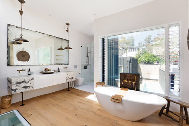 This is an example of a contemporary master bathroom in Sydney with flat-panel cabinets, white cabinets, a freestanding tub, an alcove shower, a wall-mount toilet, white tile, white walls, light hardwood floors, a drop-in sink, beige floor, a hinged shower door and white benchtops.