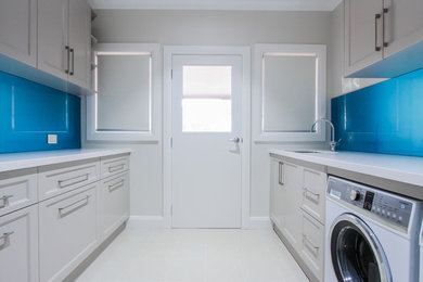 Photo of a mid-sized contemporary single-wall utility room in Sydney with an undermount sink, granite benchtops, beige walls, ceramic floors, a stacked washer and dryer, beige floor and white cabinets.