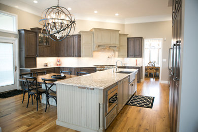 Design ideas for a large transitional u-shaped kitchen in Indianapolis with a farmhouse sink, raised-panel cabinets, dark wood cabinets, medium hardwood floors, with island, brown floor, granite benchtops, white splashback, subway tile splashback, panelled appliances and multi-coloured benchtop.