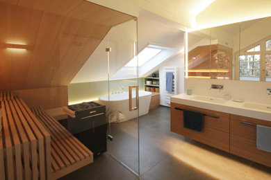 Großes Modernes Badezimmer En Suite mit flächenbündigen Schrankfronten, hellen Holzschränken, freistehender Badewanne, Nasszelle, Wandtoilette, grauen Fliesen, Keramikfliesen, grüner Wandfarbe, Keramikboden, integriertem Waschbecken, Mineralwerkstoff-Waschtisch, grauem Boden und weißer Waschtischplatte in München