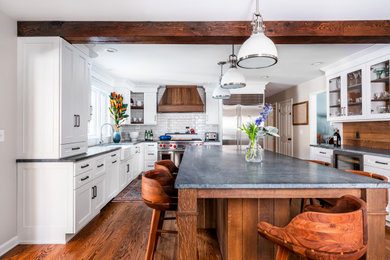 This is an example of a mid-sized country u-shaped eat-in kitchen in New York with a farmhouse sink, shaker cabinets, medium wood cabinets, soapstone benchtops, white splashback, ceramic splashback, stainless steel appliances, medium hardwood floors, with island, brown floor, grey benchtop and exposed beam.
