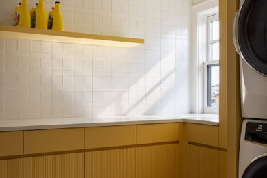 Example of a mid-sized beach style l-shaped ceramic tile and multicolored floor dedicated laundry room design in New York with flat-panel cabinets, yellow cabinets, quartzite countertops, white backsplash, ceramic backsplash, white walls, a stacked washer/dryer and white countertops