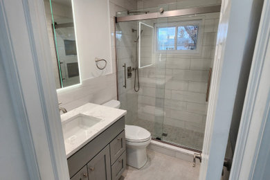 Inspiration for a contemporary bathroom in New York with recessed-panel cabinets, grey cabinets, a two-piece toilet, white tiles, porcelain tiles, white walls, porcelain flooring, a submerged sink, quartz worktops, grey floors, a sliding door, white worktops, a wall niche and a single sink.