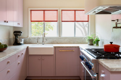 Example of a trendy u-shaped medium tone wood floor and brown floor kitchen design in Los Angeles with a farmhouse sink, flat-panel cabinets, pink cabinets, stainless steel appliances and beige countertops