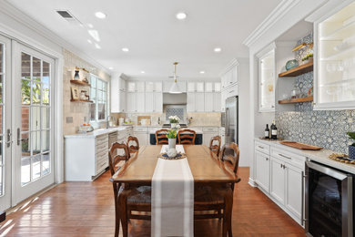 Example of a mid-sized transitional u-shaped medium tone wood floor and brown floor eat-in kitchen design in Other with a farmhouse sink, shaker cabinets, white cabinets, quartzite countertops, gray backsplash, porcelain backsplash, stainless steel appliances and an island
