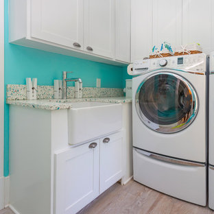 This is an example of a medium sized beach style galley utility room in Charleston with a belfast sink, raised-panel cabinets, white cabinets, recycled glass countertops, blue walls, light hardwood flooring, a side by side washer and dryer, beige floors and multicoloured worktops.