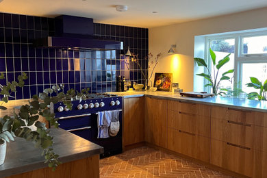 Photo of a medium sized bohemian u-shaped kitchen/diner in Sussex with a belfast sink, flat-panel cabinets, light wood cabinets, blue splashback, porcelain splashback, stainless steel appliances, terracotta flooring, no island, orange floors, grey worktops and a feature wall.