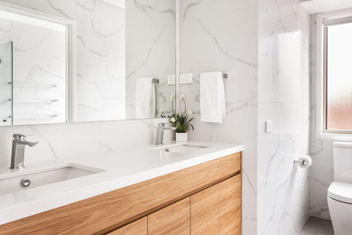 This is an example of a mid-sized contemporary master bathroom in Sydney with recessed-panel cabinets, brown cabinets, a drop-in tub, a corner shower, a one-piece toilet, white tile, marble, white walls, cement tiles, an undermount sink, solid surface benchtops, grey floor, a hinged shower door and white benchtops.