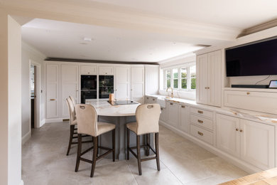 Large farmhouse grey and white l-shaped open plan kitchen in Essex with a belfast sink, shaker cabinets, grey cabinets, engineered stone countertops, black appliances, ceramic flooring, an island, beige floors and white worktops.