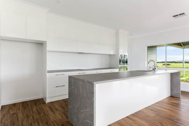 Photo of a mid-sized country galley open plan kitchen in Adelaide with a drop-in sink, flat-panel cabinets, white cabinets, laminate benchtops, white splashback, porcelain splashback, stainless steel appliances, laminate floors, with island, brown floor and grey benchtop.