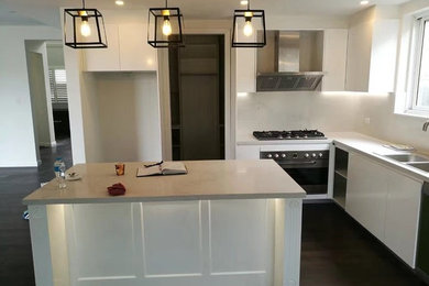 This is an example of a mid-sized modern l-shaped kitchen in Sydney with a drop-in sink, white cabinets, quartz benchtops, white splashback, stainless steel appliances, laminate floors, with island and brown floor.