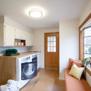 フィラデルフィアの中サイズのトランジショナルスタイルのおしゃれな家事室 (アンダーカウンターシンク、シェーカースタイル扉のキャビネット、白いキャビネット、木材カウンター、白い壁、コルクフローリング、左右配置の洗濯機・乾燥機、茶色い床、ベージュのキッチンカウンター) の写真