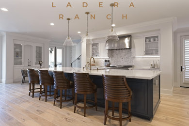 Example of a large minimalist l-shaped light wood floor and beige floor open concept kitchen design in Orange County with an undermount sink, shaker cabinets, white cabinets, quartz countertops, white backsplash, porcelain backsplash, stainless steel appliances, an island and white countertops