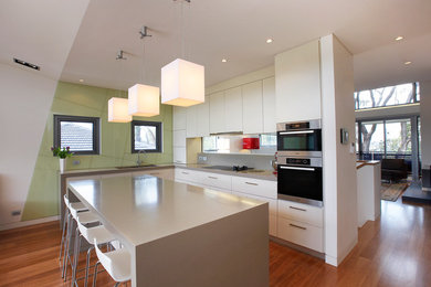 Design ideas for a mid-sized contemporary galley eat-in kitchen in Sydney with a double-bowl sink, flat-panel cabinets, white cabinets, quartz benchtops, green splashback, glass sheet splashback, stainless steel appliances, medium hardwood floors, with island, red floor, beige benchtop and vaulted.