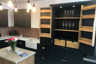 Medium sized contemporary l-shaped kitchen/diner in London with a belfast sink, beaded cabinets, blue cabinets, wood worktops, glass sheet splashback, black appliances, porcelain flooring, an island, white floors and brown worktops.