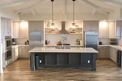 Mid-sized transitional u-shaped kitchen in Hawaii with a farmhouse sink, raised-panel cabinets, white cabinets, solid surface benchtops, white splashback, subway tile splashback, stainless steel appliances, medium hardwood floors, with island, brown floor and white benchtop.