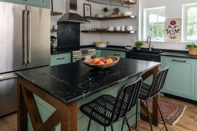 Small farmhouse l-shaped laminate floor and brown floor enclosed kitchen photo in Philadelphia with a farmhouse sink, shaker cabinets, green cabinets, soapstone countertops, black backsplash, stone slab backsplash, stainless steel appliances, an island and black countertops