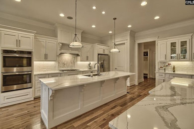 Example of a transitional medium tone wood floor and brown floor kitchen design in Charlotte with an undermount sink, shaker cabinets, white cabinets, quartz countertops, white backsplash, ceramic backsplash, stainless steel appliances and an island