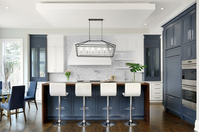 Design ideas for a large transitional l-shaped open plan kitchen in Toronto with recessed-panel cabinets, blue cabinets, wood benchtops, white splashback, dark hardwood floors, with island, brown floor, a drop-in sink, marble splashback and stainless steel appliances.