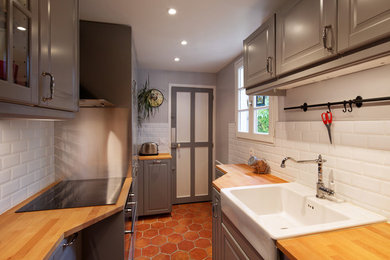 This is an example of a small contemporary galley separate kitchen in Paris with a farmhouse sink, flat-panel cabinets, grey cabinets, wood benchtops, white splashback, subway tile splashback, panelled appliances, terra-cotta floors and no island.