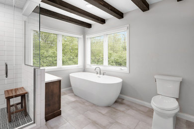 This is an example of a large modern master bathroom in Detroit with shaker cabinets, medium wood cabinets, a freestanding tub, an alcove shower, a two-piece toilet, white tile, porcelain tile, beige walls, porcelain floors, an undermount sink, engineered quartz benchtops, beige floor, a hinged shower door, white benchtops, a niche, a double vanity, a freestanding vanity and exposed beam.