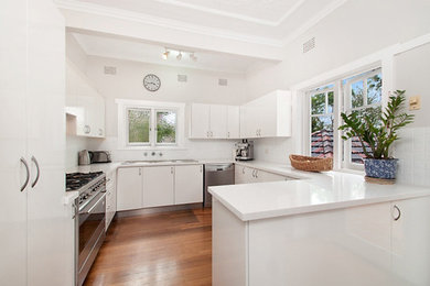 Photo of a contemporary u-shaped kitchen in Sydney with flat-panel cabinets, white cabinets, white splashback, stainless steel appliances, medium hardwood floors and a peninsula.