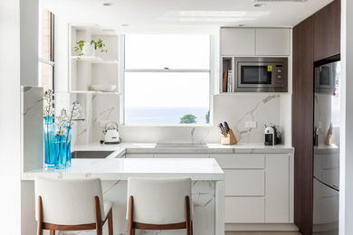 This is an example of a beach style u-shaped kitchen in Sydney with a drop-in sink, flat-panel cabinets, white cabinets, white splashback, stainless steel appliances, beige floor and white benchtop.