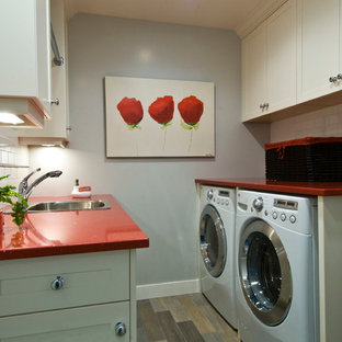 バンクーバーの中サイズのトラディショナルスタイルのおしゃれな洗濯室 (ドロップインシンク、シェーカースタイル扉のキャビネット、白いキャビネット、クオーツストーンカウンター、グレーの壁、磁器タイルの床、左右配置の洗濯機・乾燥機、赤いキッチンカウンター) の写真