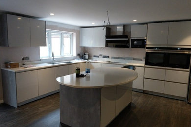 Inspiration for a large contemporary l-shaped open plan kitchen in Hertfordshire with a double-bowl sink, flat-panel cabinets, grey cabinets, composite countertops, black appliances, dark hardwood flooring, an island, brown floors and white worktops.
