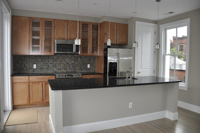This is an example of a mid-sized contemporary single-wall eat-in kitchen in DC Metro with a drop-in sink, glass-front cabinets, medium wood cabinets, laminate benchtops, multi-coloured splashback, stone tile splashback, stainless steel appliances, light hardwood floors, with island, beige floor and black benchtop.