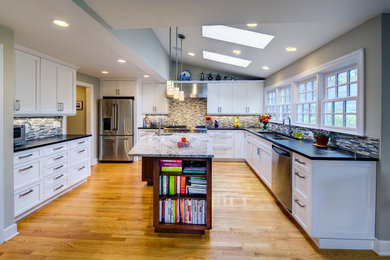 Mid-sized elegant l-shaped medium tone wood floor open concept kitchen photo in Detroit with an undermount sink, recessed-panel cabinets, white cabinets, soapstone countertops, gray backsplash, ceramic backsplash, stainless steel appliances and an island