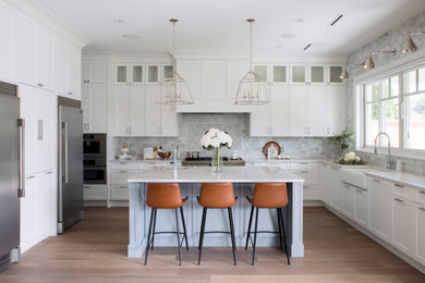 Transitional u-shaped kitchen in Vancouver with a farmhouse sink, shaker cabinets, white cabinets, grey splashback, stainless steel appliances, medium hardwood floors, with island, brown floor, white benchtop, quartz benchtops and marble splashback.