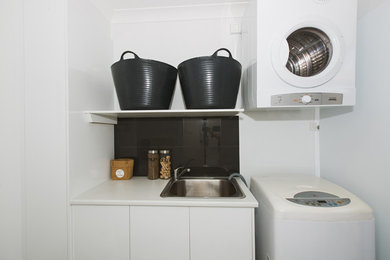 This is an example of a modern galley dedicated laundry room in Sydney with a single-bowl sink, white cabinets, laminate benchtops, white walls, ceramic floors and a stacked washer and dryer.